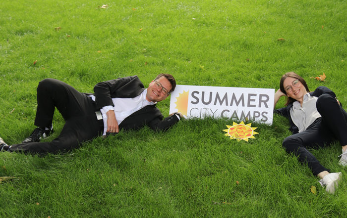 Julia Herr und Jürgen Czernohorszky liegen in der Wiese mit einem Schild der Summer City Camps